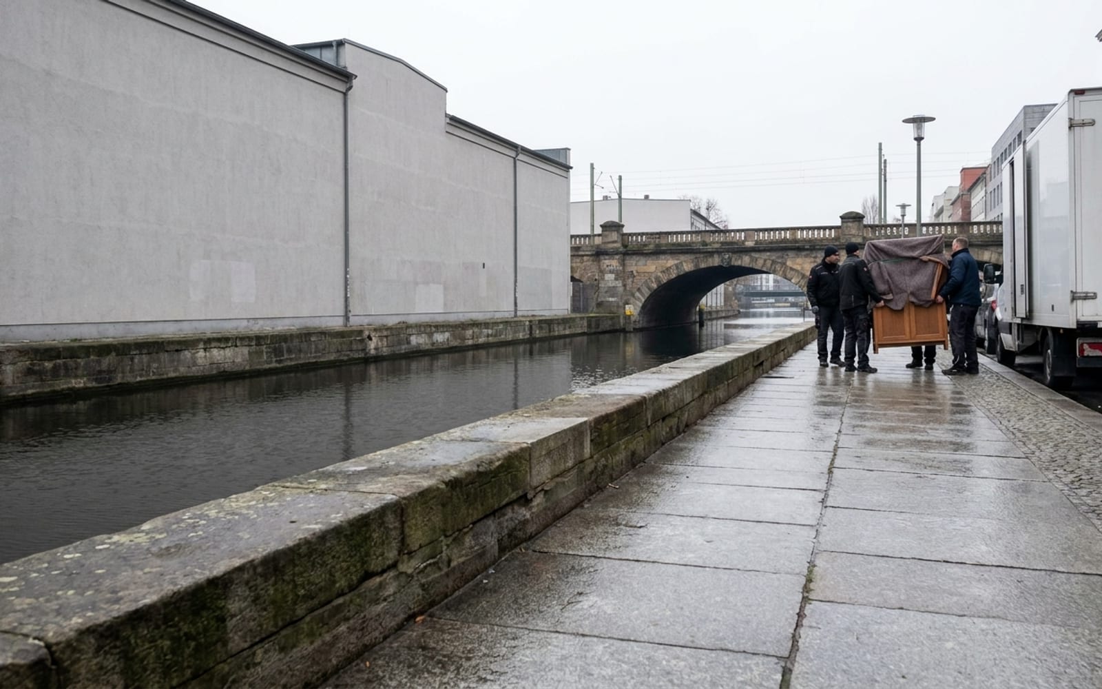 Ruhige gepflasterte Uferpromenade an einem Kanal, rechts hinten ein Umzugswagen von der Seite und drei Helfer mit einem eingepackten Schrank vor einer steinernen Bogenbrücke