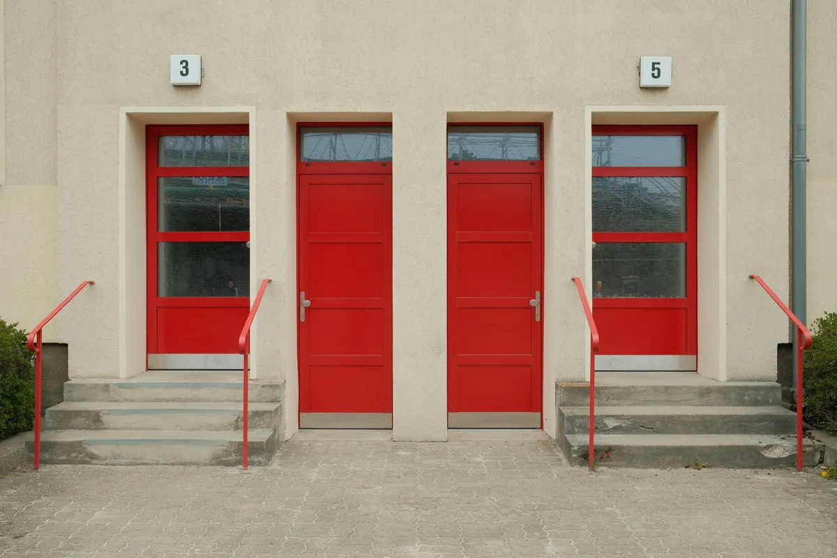 Zwei rote Hauseingangstueren mit den Nummern 3 und 5 an einem schlichten Putzbau, davor kurze Treppen mit roten Handläufen